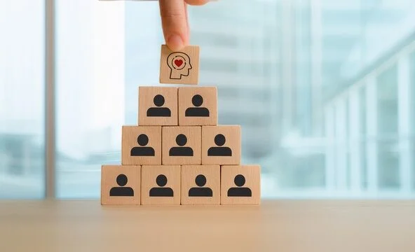 Une main place un bloc de bois avec une tête humaine et un cœur au-dessus d’une pyramide de blocs avec des icônes humaines, symbolisant le leadership, l’intelligence émotionnelle et la hiérarchie organisationnelle.