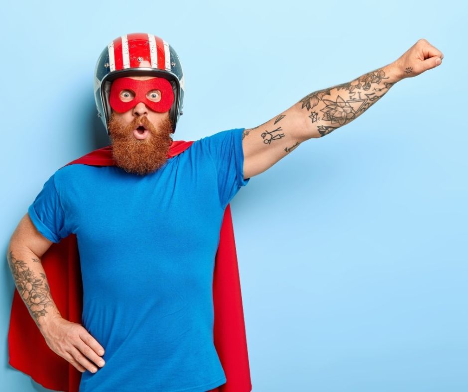 Un homme barbu portant une cape rouge, un masque rouge et un casque pose avec un bras levé, prêt à conquérir les clics et à maîtriser le marketing numérique, sur un fond bleu clair.