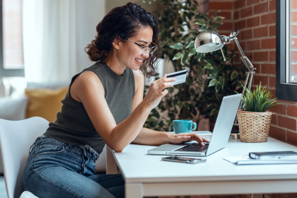Femme assise à un bureau, tenant une carte de crédit tout en utilisant un ordinateur portable, entourée d'une tasse, d'un téléphone et d'une plante en pot. Cette image illustre parfaitement le monde du marketing numérique et des clics en ligne sans interruption.