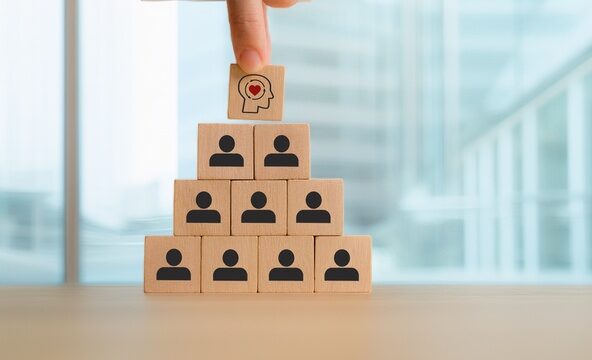 Une main place un bloc de bois avec une tête humaine et un cœur au-dessus d'une pyramide de blocs avec des icônes humaines, symbolisant le leadership, l'intelligence émotionnelle et la hiérarchie organisationnelle.