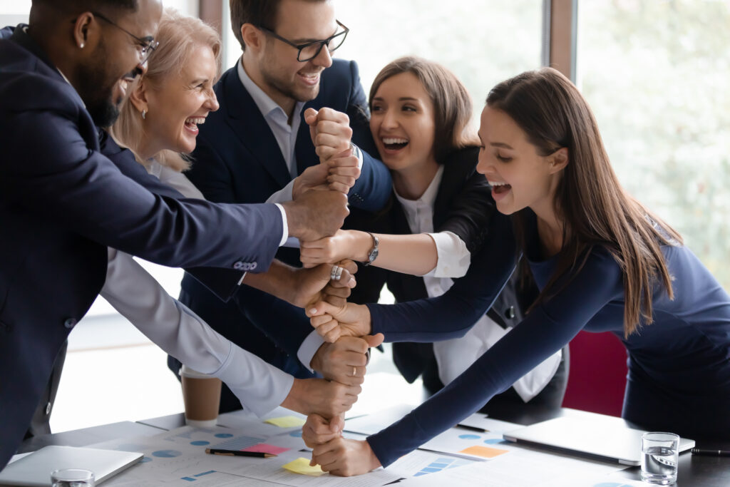 Un groupe de cinq personnes habillées professionnellement se tient autour d'une table, empilant leurs mains et souriant dans un environnement de bureau lumineux, mettant en évidence leur forte intelligence émotionnelle et leur esprit d'équipe.