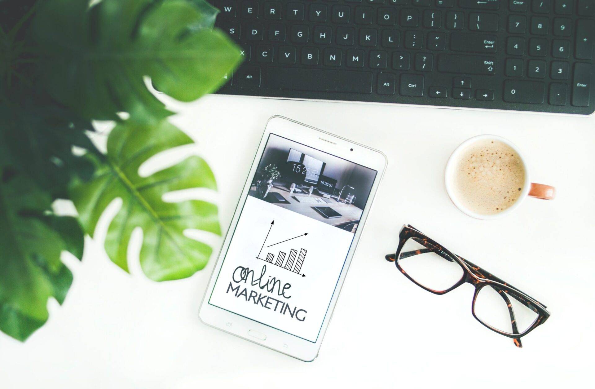 Un smartphone affichant « marketing en ligne » sur son écran, placé à côté d'une tasse de café, de lunettes et d'un clavier sur un bureau blanc avec une plante verte et des notes sur « avenir