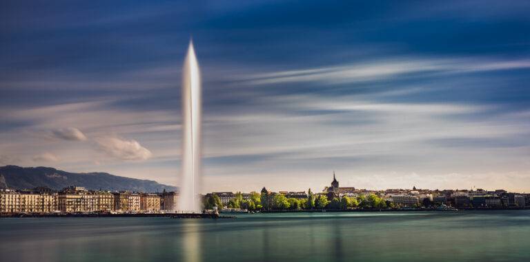 A towering water jet dominates the city skyline by a lake.