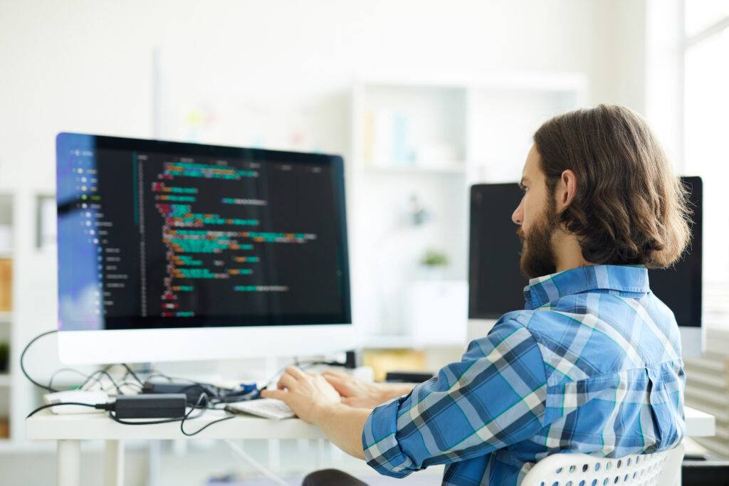 Man with beard coding on computer in bright office.